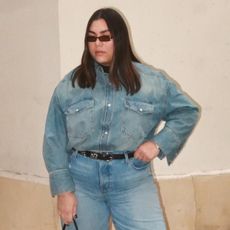 Woman wears denim shirt, denim jeans, and black belt with sunglasses standing in front of cream-colored wall.