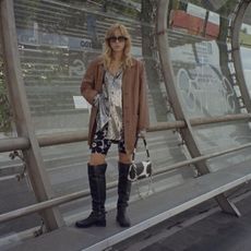 a casual party outfit shown on a woman standing at a bus stop wearing a tan blazer over a sequin button-down shirt with printed capri pants and over-the-knee moto boots