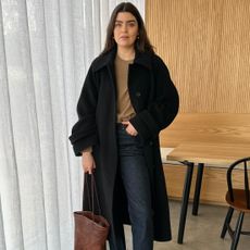 British fashion influencer Anna Newton poses in her modern wood paneled kitchen in London wearing a winter outfit with an oversize black coat, a camel crewneck sweater, dark wash jeans, and a woven brown leather tote bag