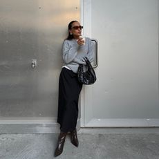 Fashion influencer @ingridedvinsen wearing an elegant everyday outfit with a black skirt, knee-high boots, a woven leather bag, grey sweater worn over a white t-shirt, and sunglasses.