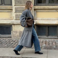 Danish art director Clara Dyrhauge walking on a cobblestone sidewalk with effortless hair wearing an oversize gray belted coat, a brown suede bag, straight-leg jeans, and black kitten-heel boots